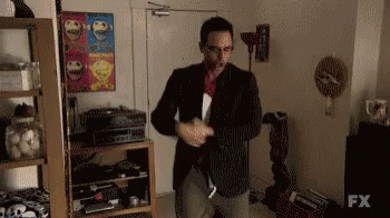 A Man In A Suit Is Standing In A Living Room With A Sign That Says Fx On It . GIF