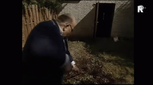 A Man In A Suit Is Standing In Front Of A Brick Building With The Letter R On The Bottom Of The Screen GIF