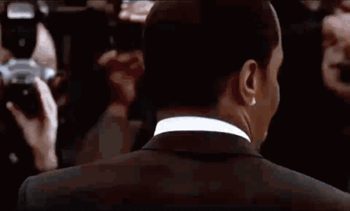 A Man In A Suit Is Standing In Front Of A Crowd Of People And A Camera . GIF