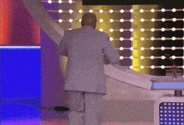 A Man In A Suit Is Standing In Front Of A Stage With Lights Behind Him GIF