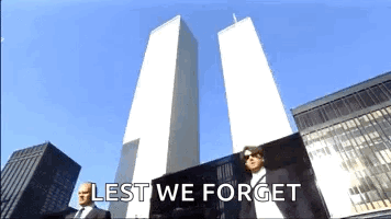 A Man In A Suit Is Standing In Front Of The Twin Towers With The Words Lest We Forget Written Below Him GIF