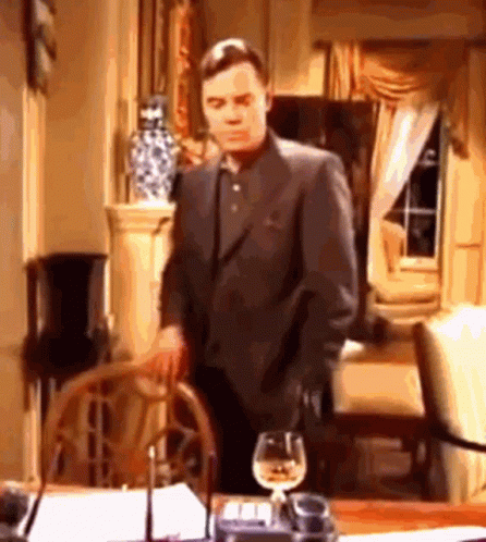 A Man In A Suit Is Standing Next To A Table With A Glass Of Wine . GIF