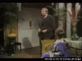 A Man In A Suit Is Standing Next To A Woman In A Purple Dress Who Is Sitting In A Chair GIF