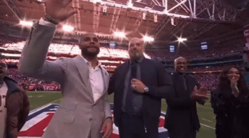 A Man In A Suit Is Standing On A Football Field With A Group Of People Applauding . GIF