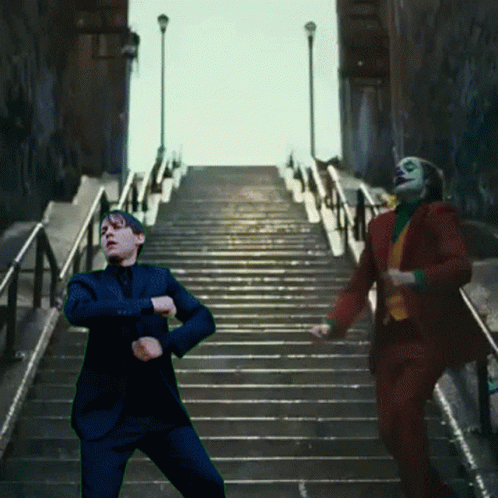 A Man In A Suit Is Standing On A Set Of Stairs Next To A Man In A Clown Suit GIF