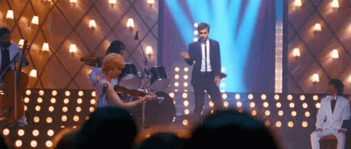 A Man In A Suit Is Standing On A Stage With A Woman Playing A Violin . GIF