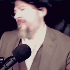 A Man In A Suit Is Talking Into A Microphone While Wearing A Hat . GIF