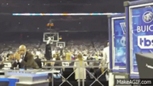 a man in a suit is talking on a cell phone in front of a crowd at a basketball game sponsored by buick .