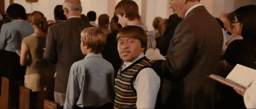 A Man In A Suit Is Talking To A Boy In A Striped Vest In A Church GIF