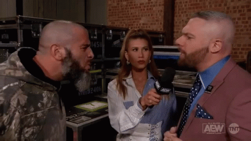 A Man In A Suit Is Talking To A Woman In A Microphone While Another Man Looks On . GIF