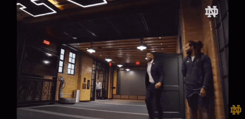 A Man In A Suit Is Walking In A Room With A Sign That Says Exit On It GIF
