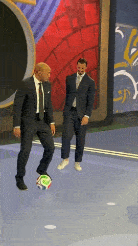 A Man In A Suit Kicks A Soccer Ball While Another Man Stands Behind Him GIF