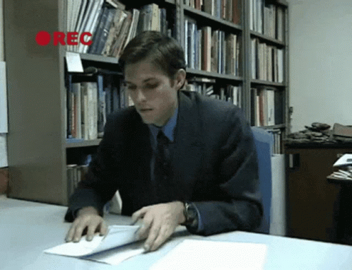 A Man In A Suit Sits At A Desk In Front Of A Bookshelf With The Word Rec On It GIF