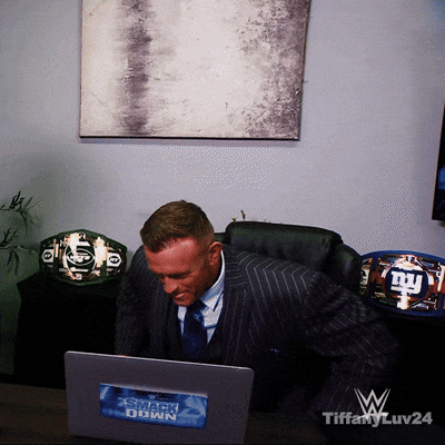 A Man In A Suit Sits At A Desk In Front Of A Laptop With A Sticker On It That Says Smackdown GIF
