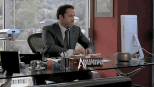 A Man In A Suit Sits At A Desk With A Sign That Says Atlantis On It GIF