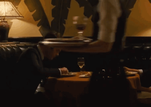 A Man In A Suit Sits At A Table While A Waiter Holds A Tray Of Food GIF