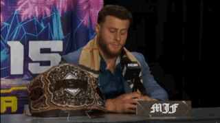 A Man In A Suit Sits At A Table With A Microphone And A Championship Belt On It GIF