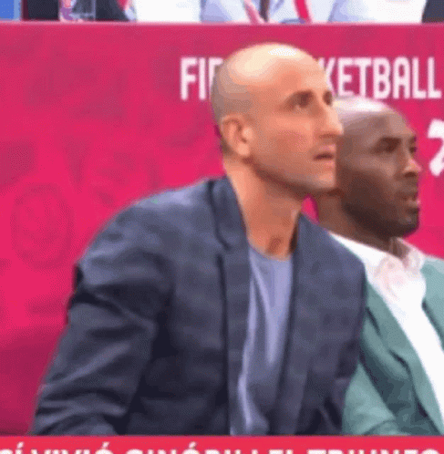A Man In A Suit Sits In Front Of A Red Sign That Says Basketball GIF