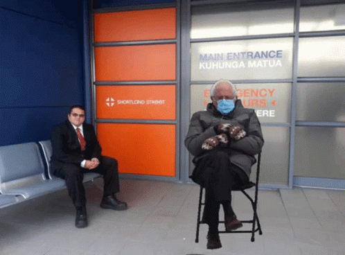 A Man In A Suit Sits Next To A Man In A Chair In Front Of A Sign That Says Main Entrance GIF