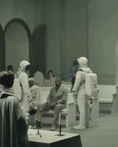 A Man In A Suit Sits On A Bench While A Group Of Astronauts Stand Around Him GIF
