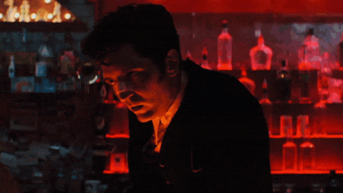 A Man In A Suit Stands In Front Of A Bar With Bottles Of Alcohol On Shelves GIF