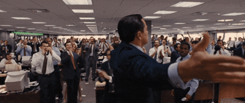 A Man In A Suit Stands In Front Of A Crowd Of People In An Office With A Sign On The Wall That Says " Stocks " GIF