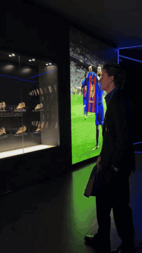 A Man In A Suit Stands In Front Of A Display Case With Soccer Trophies And A Shirt That Says Messi GIF