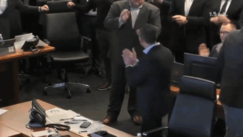 A Man In A Suit Stands In Front Of A Group Of People Applauding With The Letters Ts On The Bottom Left GIF