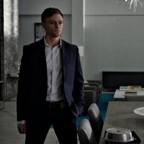 A Man In A Suit Stands In Front Of A Table With His Hands In His Pockets GIF