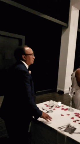 A Man In A Suit Stands In Front Of A Table With Wine Glasses And Rose Petals On It GIF