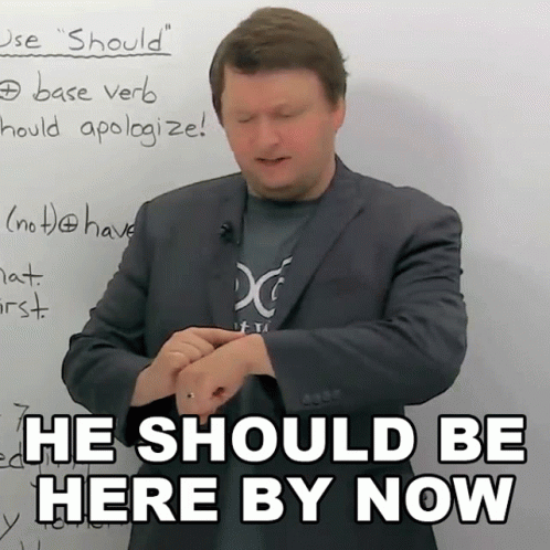 A Man In A Suit Stands In Front Of A White Board With The Words He Should Be Here By Now GIF
