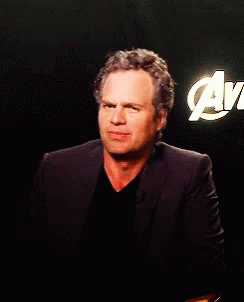 A Man In A Suit Stands In Front Of An Avengers Logo GIF