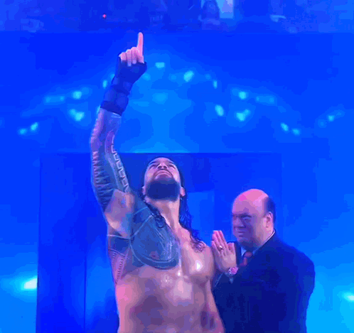 A Man In A Suit Stands Next To A Shirtless Wrestler Who Is Pointing Up At The Sky GIF
