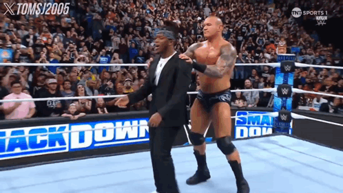 A Man In A Suit Stands Next To A Wrestler In A Wrestling Ring That Says Back Down GIF