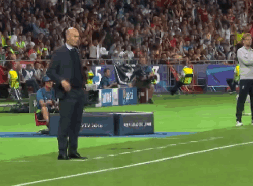 A Man In A Suit Stands On A Soccer Field In Front Of A Banner That Says 5 GIF