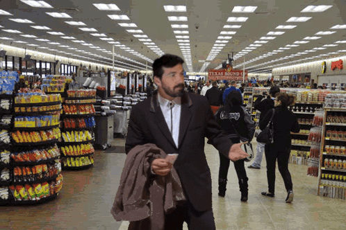 A Man In A Suit Walks Through A Grocery Store With A Sign That Says Texas Roundup GIF