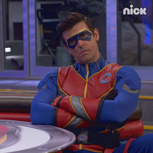 A Man In A Superhero Costume Is Sitting At A Table With His Arms Crossed And A Nick Logo Behind Him GIF