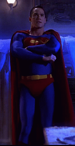 A Man In A Superman Costume Is Standing In A Room With His Arms Crossed GIF