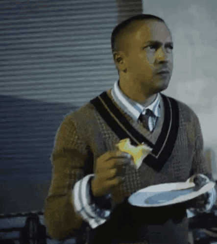 A Man In A Sweater And Tie Is Holding A Plate Of Food And Eating A Piece Of Cheese . GIF