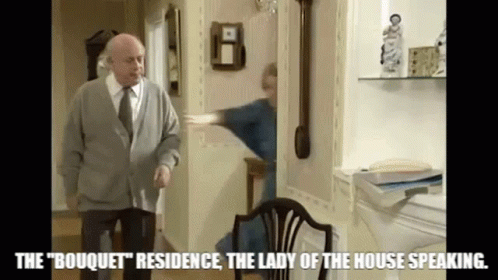 A Man In A Sweater And Tie Is Standing In A Living Room Next To A Woman In A Blue Robe . GIF