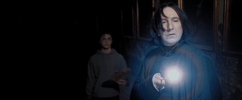 A Man In A Sweater And Tie Stands In A Dark Room With A Light Shining On Him GIF