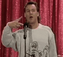 A Man In A Sweater Is Giving A Speech In Front Of A Microphone And Pointing At The Camera . GIF