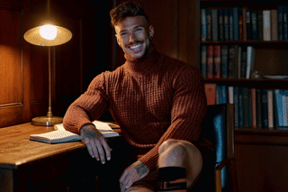 A Man In A Sweater Is Sitting At A Desk With A Lamp GIF