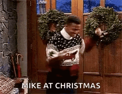 A Man In A Sweater Is Standing In Front Of A Door Holding A Wreath And Says `` Mike At Christmas '' GIF