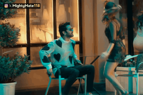 A Man In A Sweater With Hearts On It Sits Next To A Woman In A Short Dress GIF