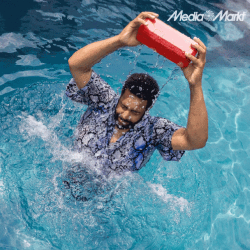 a man in a swimming pool is holding a red box over his head with the media markt logo in the corner