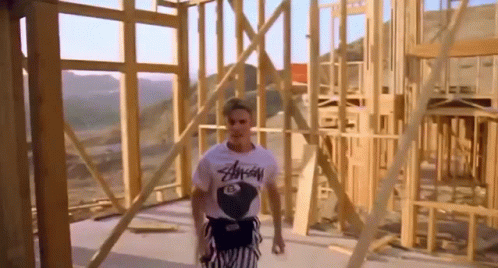 A Man In A T-Shirt Is Standing In Front Of A Wooden Structure Under Construction . GIF