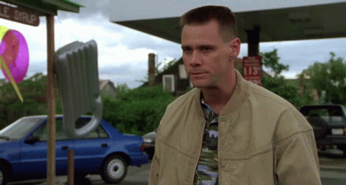 A Man In A Tan Jacket Stands In Front Of A Gas Station With A Sign That Says 113 GIF