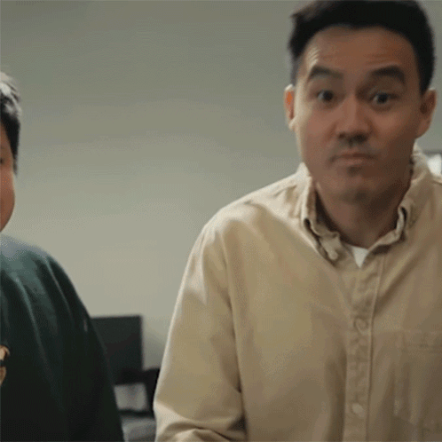 A Man In A Tan Shirt Is Making A Funny Face While Standing Next To Another Man In A Green Sweater . GIF