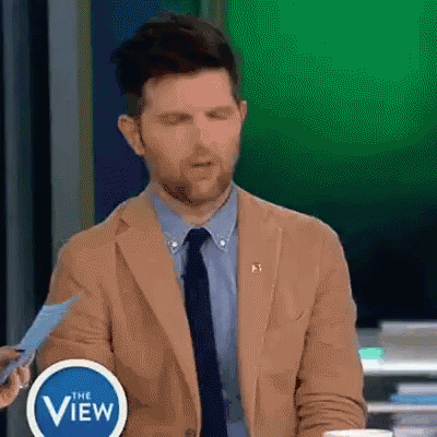 A Man In A Tan Suit And Tie Is Holding A Piece Of Paper In Front Of A Sign That Says The View . GIF